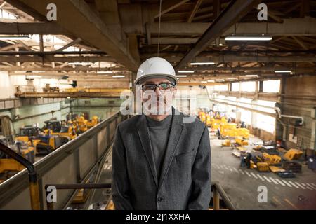 Portrait of senior architect in work helmet looking at camera while standing on balcony in plant with production equipment in the background Stock Photo