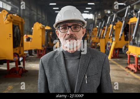 Portrait of senior engineer in work helmet looking at camera while standing in warehouse with new machines in the background Stock Photo