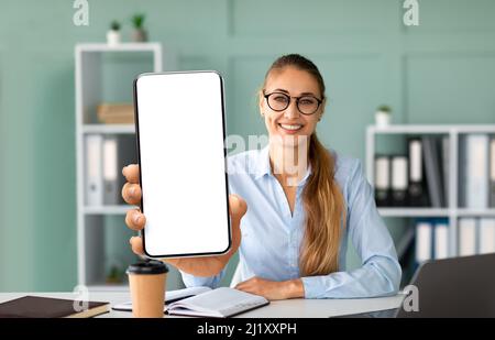 Positive businesswoman showing modern cellphone with big blank screen, sitting at workplace in office, mockup Stock Photo