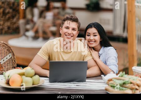 Portrait of happy young multiracial couple with laptop sitting at table near camper van, smiling at camera outdoors Stock Photo
