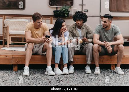 Group of diverse friends sitting near camper van, speaking to each other, using smartphones on camping trip Stock Photo