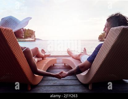 Just the two of us. Rearview shot of a cheerful young couple holding hands while relaxing on deck chairs and looking out at the ocean outside during Stock Photo