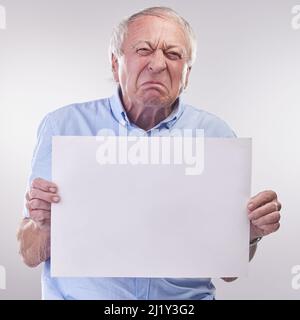 Who are you calling a grumpy old man. Studio shot of a senior man holding a blank sign and looking unhappy against a grey background. Stock Photo