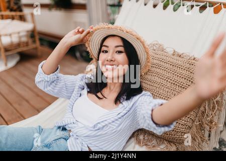 Smiling young Asian woman lying in hammock near camper van, taking selfie on camping trip in countryside Stock Photo