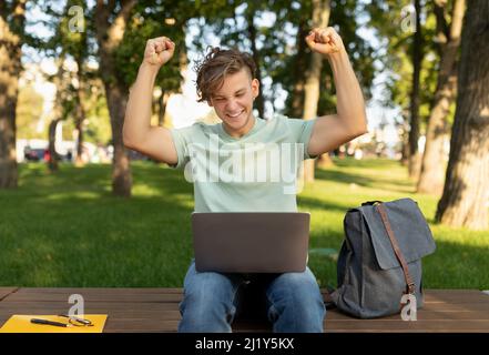 Excited student guy with laptop computer gesturing YES and celebrating success outdoors, sitting in university campus Stock Photo