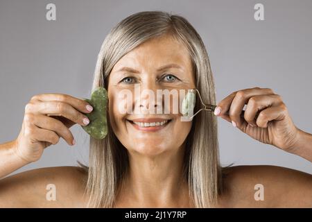 Portrait of mature caucasian woman holding gua sha massage tools while posing over grey studio background. Lady with sincere smile, blue eyes, grey hair and natural wrinkles. Stock Photo