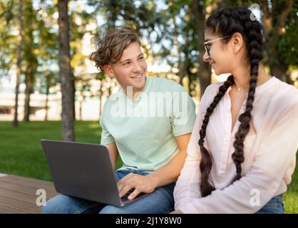 Excited students lady and guy studying together in park, preparing for exam and using laptop, sitting in park Stock Photo