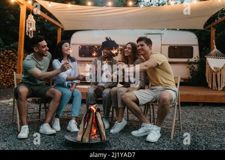 Millennial diverse friends sitting near bonfire with lit sparklers, having fun, celebrating something together on camping trip Stock Photo