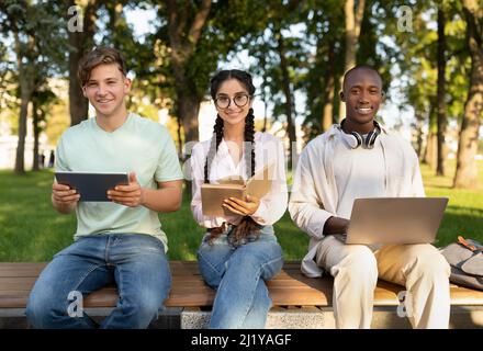 Preparing for lectures. Happy multiracial students studying together outdoors, using laptop, digital tablet and book Stock Photo
