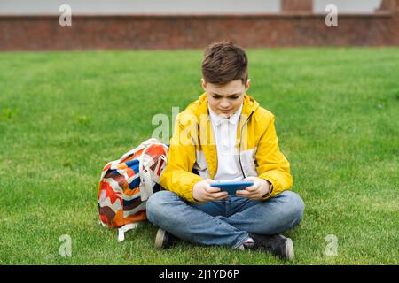 Boy playing on a smartphone sitting on a green lawn after school Stock Photo