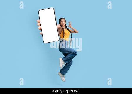 Excited young arab woman demontrating empty smartphone screen while jumping up on blue studio background, happy middle eastern female recommending new Stock Photo