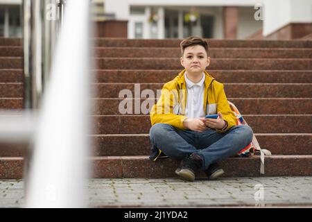 Stylishly dressed teenager sits on the stairs near the school with a smartphone in his hand Stock Photo