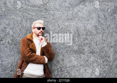 bearded hipster man with sunglasses thinking with his hand to his chin, standing against the backdrop of a building wall, copy space Stock Photo