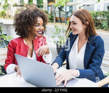 Two business women analyzing documents on laptop while sitting on a table in outdoors workplace. African woman executives at work showing some achieve Stock Photo