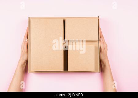 Top view to stack of cardboard boxes in female hands. Blank brown parcel boxes on pastel pink background. Packaging, shopping, free shipping, delivery Stock Photo
