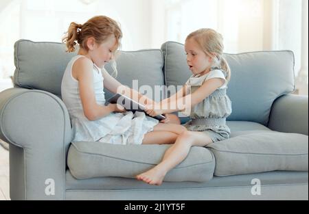 Its always your turn. Shot of two little girls fighting over a digital tablet on a sofa at home. Stock Photo