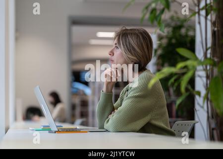 Female mature student taking break from online learning while sitting in university library Stock Photo
