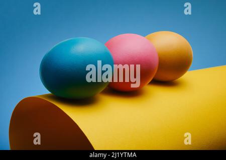 Colorful three Easter eggs on a rolled sheet of paper against a blue background Stock Photo