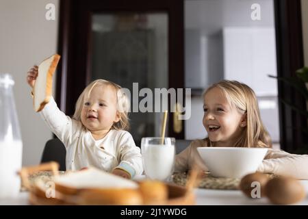 Cute caucasian siblings sitting at table on kitchen early morning and preparing breakfast with colorful cornflakes and milk. Kids enjoying life with Stock Photo