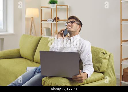 Happy young businessman talking on smartphone while sitting on couch with laptop Stock Photo