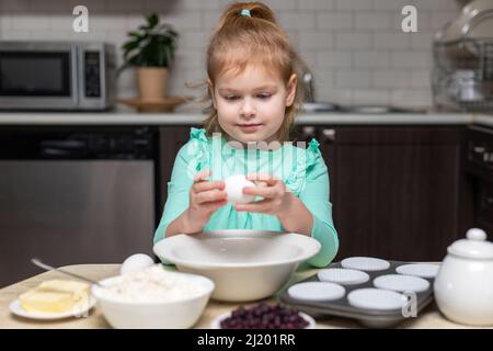 Child cooking muffins in the kitchen. Ingredients for baking on table. Little cute girl ready to break an egg to make pastry Stock Photo
