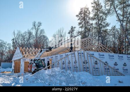 NISSWA, MN - 23 NOV 2021: Workers install new wood roof trusses on a new home construction site with winter snow. Trees line the back of the building Stock Photo