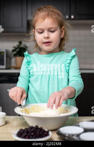 Child cooking muffins in the kitchen. Ingredients for baking on table. Little girl making dough, learning to cook at home. Stock Photo