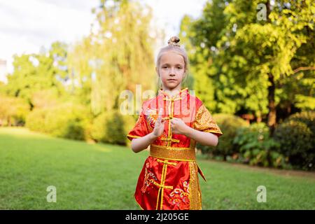 Cute little caucasian girl seven years old in red sport wushu uniform exercising in park at summer day. Lifestyle portrait of kung fu fighter child Stock Photo