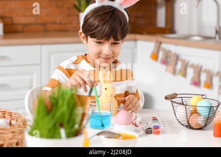 Cute little boy with figurine of bunny during painting of Easter eggs in kitchen at home Stock Photo