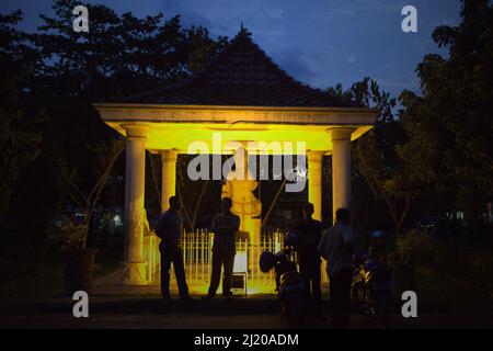 Men having conversation at dusk in front of a 7th century Srivijaya statue placed at the front yard of Sultan Mahmud Badaruddin II in Palembang, South Sumatra, Indonesia. The statue is one of the artifacts that are saved from many that remains missing or have not been found yet. According to Chau Ju-kua (Zhao Rugua), a 13th century Chinese commissioner for international trade who worked for Song Dynasty, 'There is (in San-fo-ts'i) a (kind of) Buddha (image) called 'Hill of Gold and Silver' and it is cast in gold.' Stock Photo