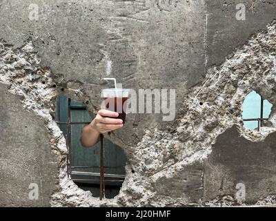 Hand holding iced coffee from the break gap of old wall. Summer drink concept. Stock Photo