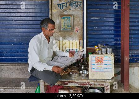 A middle-aged Indian betel (paan) vendor sits at his paan stall reading a newspaper; Mumbai, Maharashtra, India Stock Photo