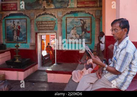 A middle-aged Indian man sits on the front porch of Chandramouleeshwara Temple in Udupi (Udipi) Karnataka, South India, reading a magazine Stock Photo