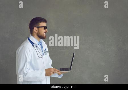 Smiling doctor in white coat working using laptop side view shot on grey wall Stock Photo
