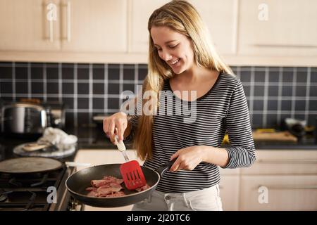 Nothing says breakfast like fried bacon. Shot of a happy young woman frying bacon on the stove at home. Stock Photo