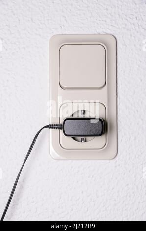 Modern light switch and german power outlet or socket on white wall indoors with black phone charger plugged in, Germany, Europe Stock Photo