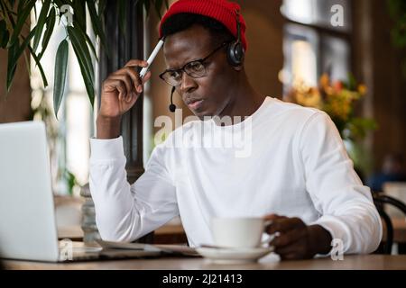 African American man in headphones sitting at cafe table taking course of E-learning, work on laptop Stock Photo