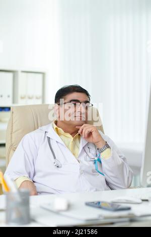 Serious doctor leaning back in his armchair when reading document on computer screen Stock Photo