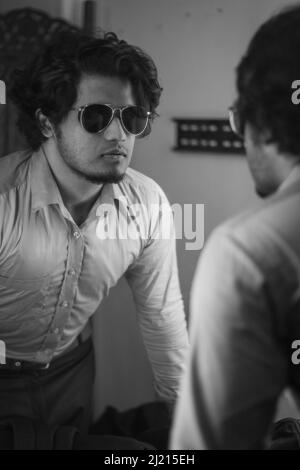 A vertical grayscale shot of an Indian man with glasses looking at his reflection in the mirror Stock Photo