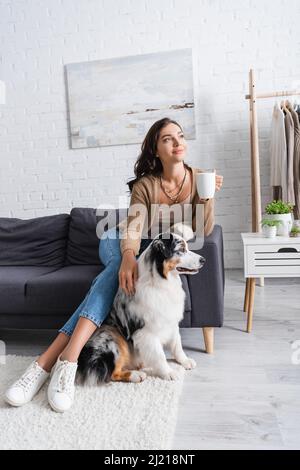 smiling young woman sitting on sofa and cuddling australian shepherd dog while holding cup of coffee Stock Photo