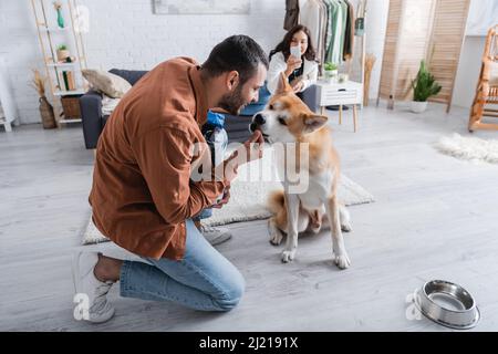 happy man feeding akita inu dog with pet food near blurred girlfriend with smartphone