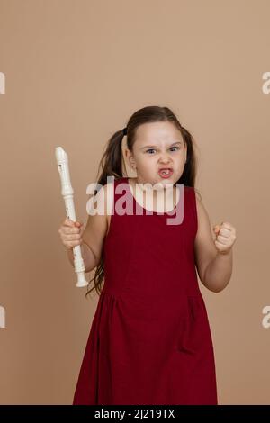Girl in red dress with angry facial expression hold flute tightly in fist, beige background. Learning to play woodwind musical instrument. Stock Photo