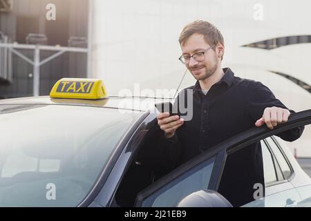 Portrait of smiling caucasian taxi driver standing near car and using smartphone. Stock Photo