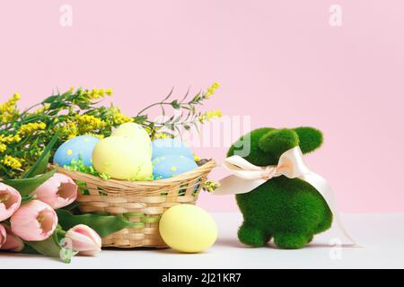 Easter composition. Basket of eggs toy rabbit flowers tulips flowering branches on the table on a pink background. Stock Photo