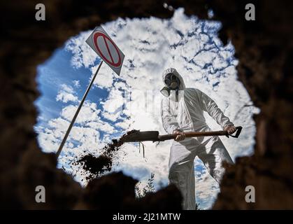 View inside pit of scientist in white suit and gas mask standing near prohibition sign and digging ground to obtain samples and study ground cover in contaminated area. Cloudy sky on the background. Stock Photo