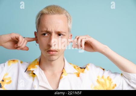 Young blonde man in shirt frowning and plugging his ears isolated over blue background Stock Photo
