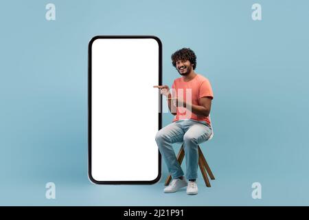 Positive indian guy in casual sitting by huge cell phone Stock Photo