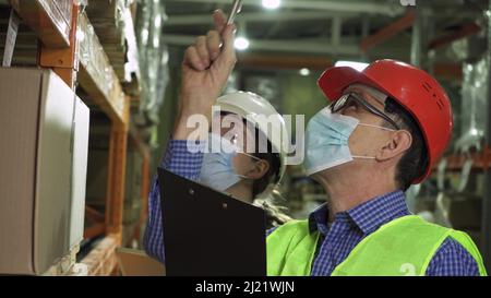 An elderly manager talks to a young female employee wearing masks and vests who works in the warehouse Stock Photo