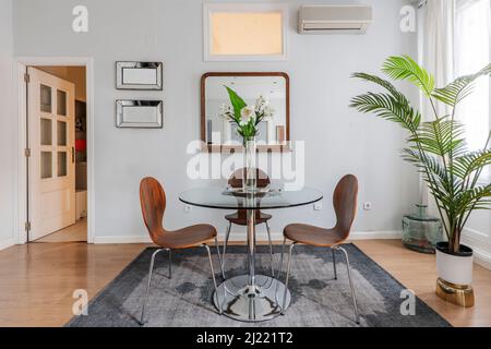 living room with round glass dining table with wooden and metal chairs, mirror on the wall and decorative palm trees in the interior Stock Photo