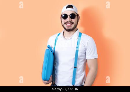 A young smiling hipster with sunglasses holding a laptop case isolated on an orange background Stock Photo